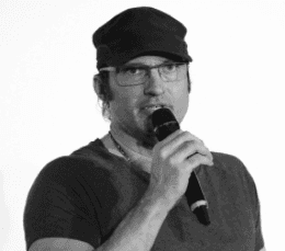 Homme portant une casquette et des lunettes, tenant un microphone devant un fond blanc
