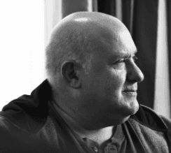 Portrait en noir et blanc d’un homme chauve regardant vers la droite, devant un rideau.