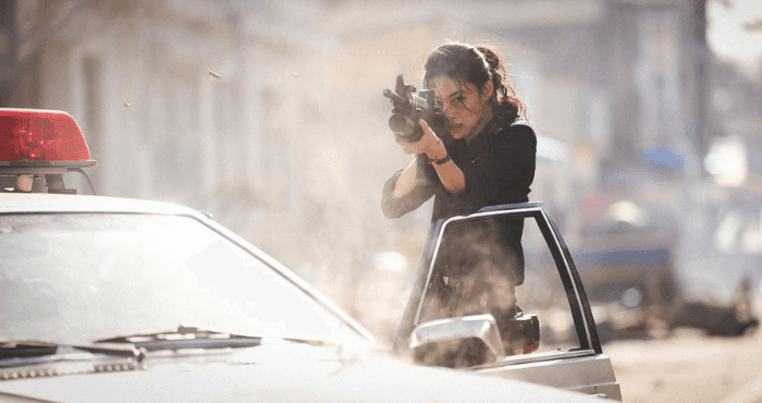 Une femme armée tire depuis la portière ouverte d'une voiture de police dans une rue urbaine.