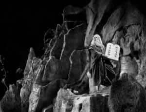 Un personnage drapé tient un livre ouvert dans une grotte rocheuse en noir et blanc