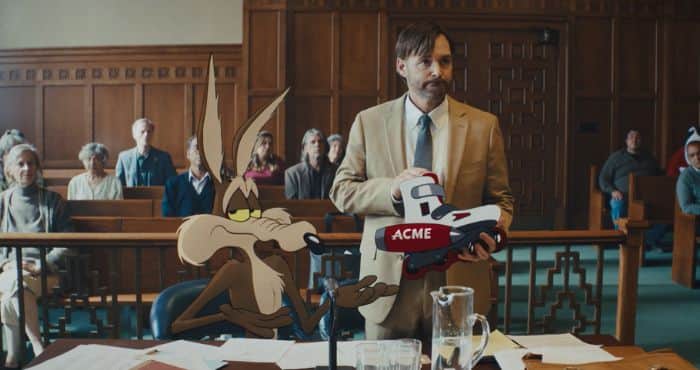 Un homme en costume tient un objet marqué « ACME » à côté d’un coyote animé dans une salle d’audience.