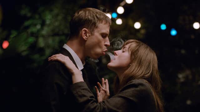 Un homme et une femme échangent de la fumée dans un jardin éclairé par des guirlandes lumineuses.