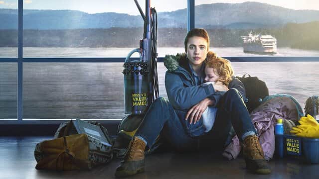 Une femme assise par terre tenant un enfant endormi, entourée de sacs et d'un aspirateur, avec un ferry en arrière-plan.