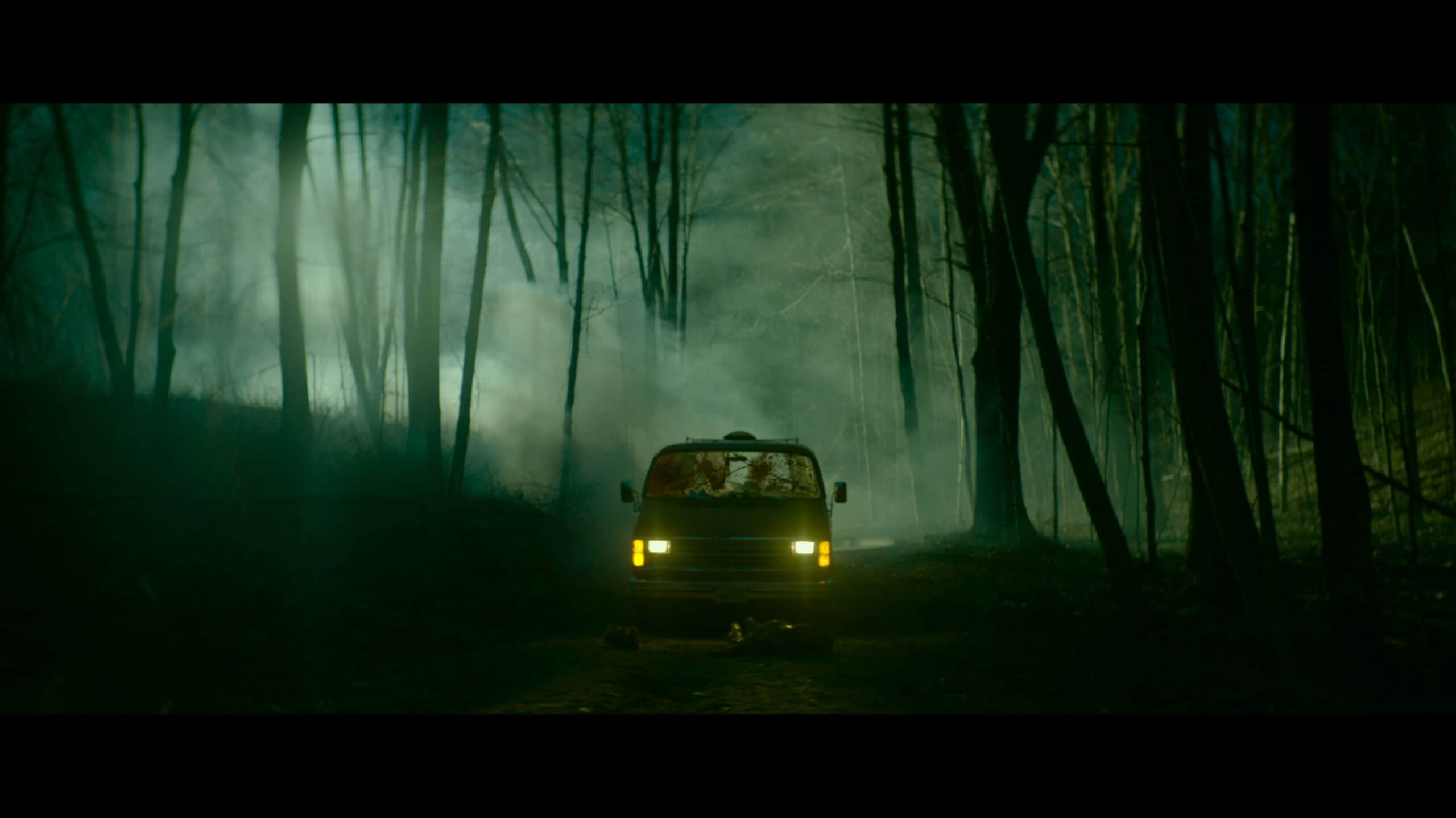 Un van aux phares allumés dans une forêt sombre et brumeuse.