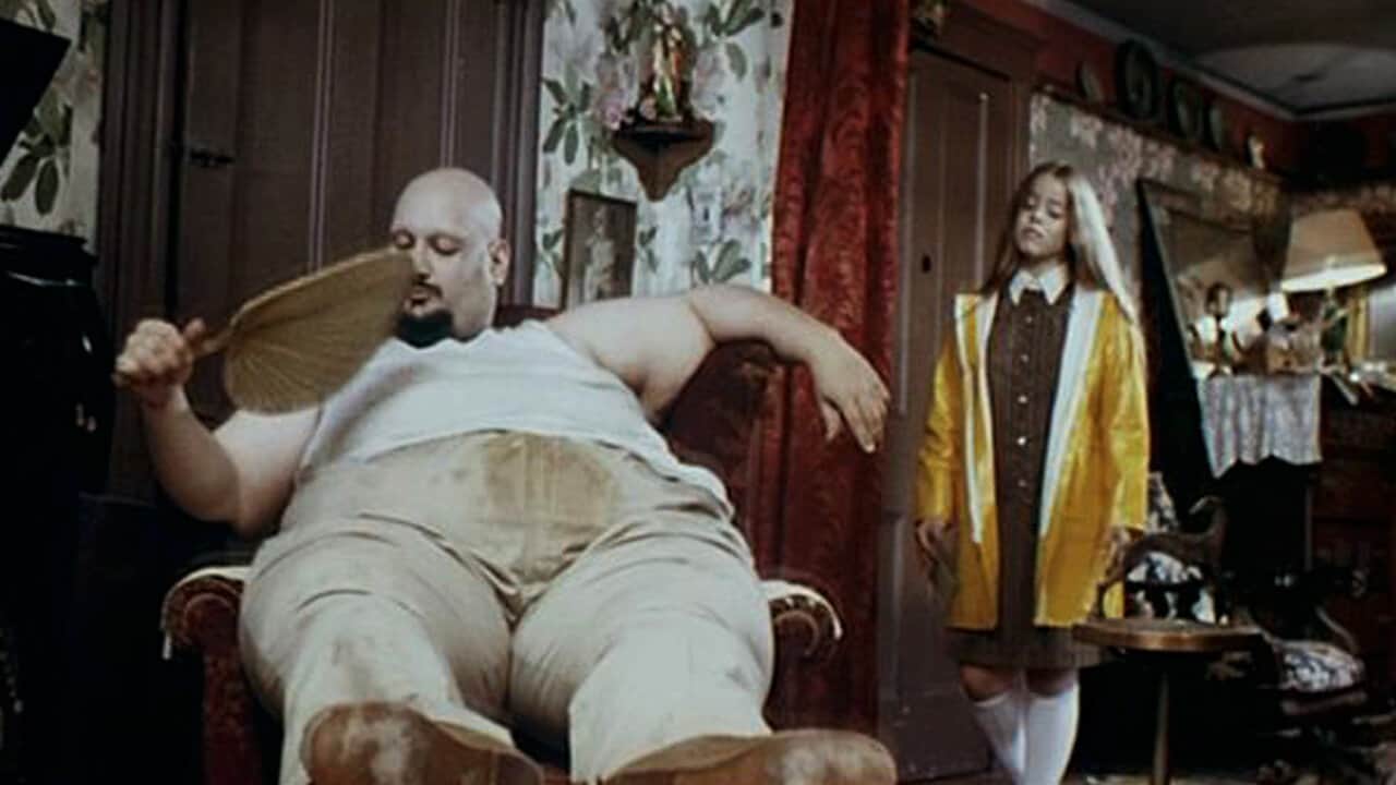 Un homme corpulent assis dans un fauteuil éventail à la main, tandis qu’une femme se tient debout dans un intérieur.