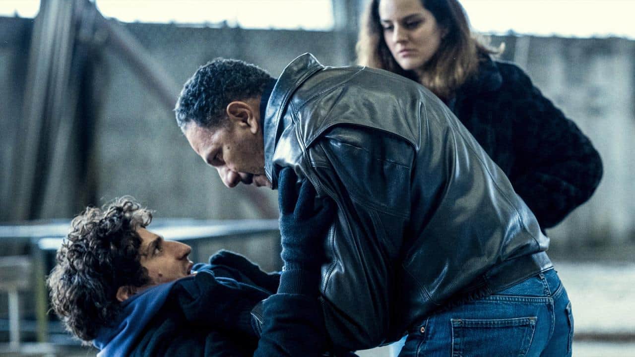 Trois personnes, dont un homme en blouson de cuir penché sur un homme allongé, sur un toit.
