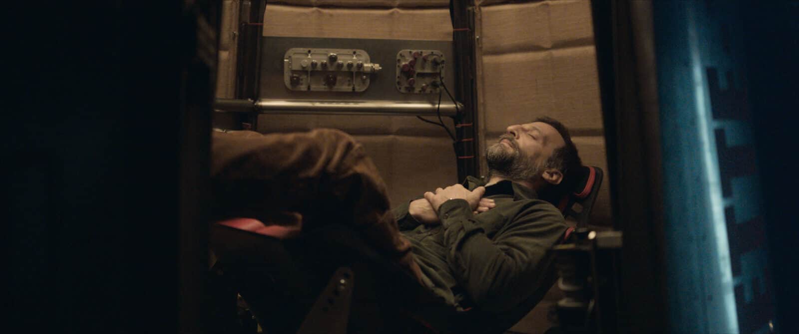 Un homme barbu allongé dans un fauteuil, les yeux fermés, dans un espace technique sombre.