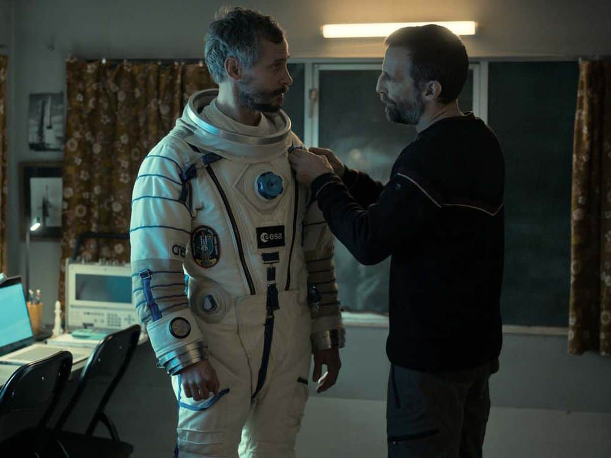 Deux hommes, dont un en combinaison spatiale, se font face dans une pièce équipée d’ordinateurs.