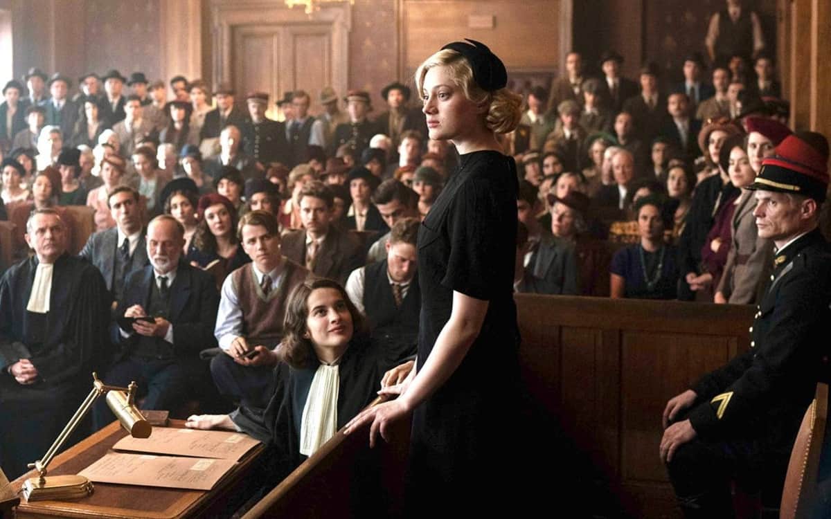 Femme debout dans une salle d’audience, face à un groupe de personnes, avec une avocate assise devant elle.
