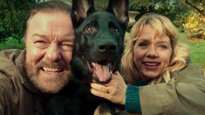 Un homme et une femme sourient avec un chien noir devant une végétation extérieure.