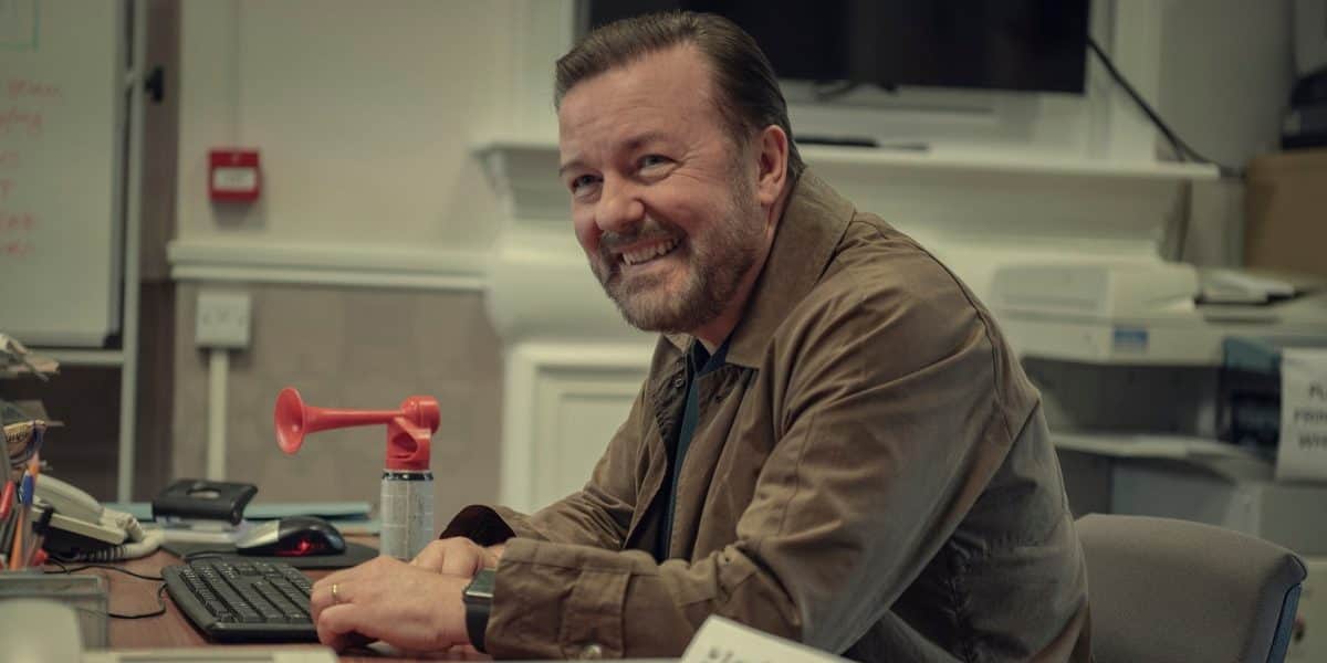 Un homme souriant assis à un bureau, devant un clavier et un klaxon rouge.