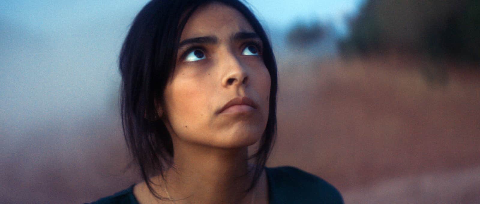 Femme aux cheveux noirs levant les yeux, dans un paysage extérieur flou au crépuscule