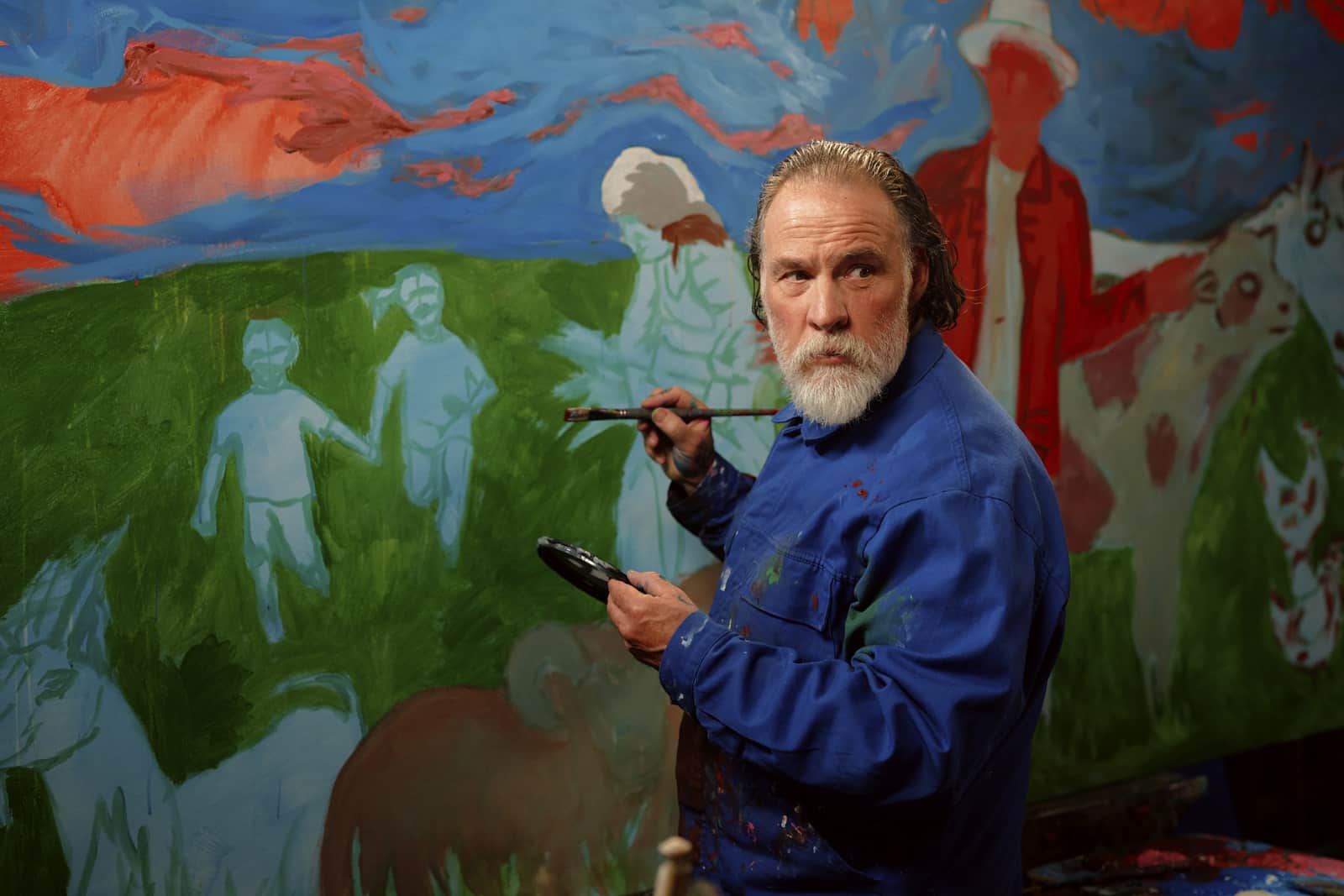 Un peintre barbu tient un pinceau et une palette devant une grande fresque colorée.