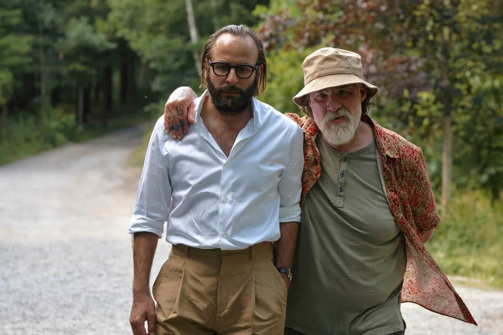 Deux hommes marchant bras dessus bras dessous sur une route forestière, l’un portant un chapeau beige.