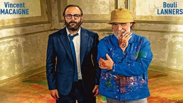 Deux hommes dans une pièce, l'un en costume et l'autre en tenue de peintre avec des mains tachées de peinture.