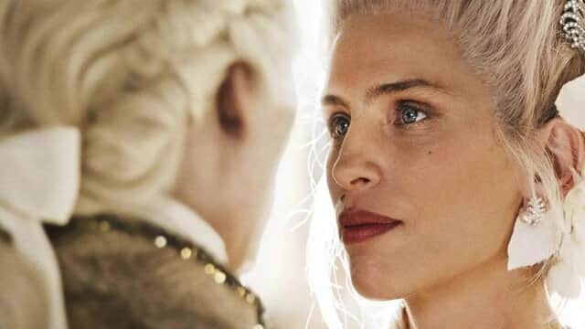 Deux personnes en costumes d'époque se regardant intensément, l'une avec des cheveux blonds et des boucles d'oreilles.