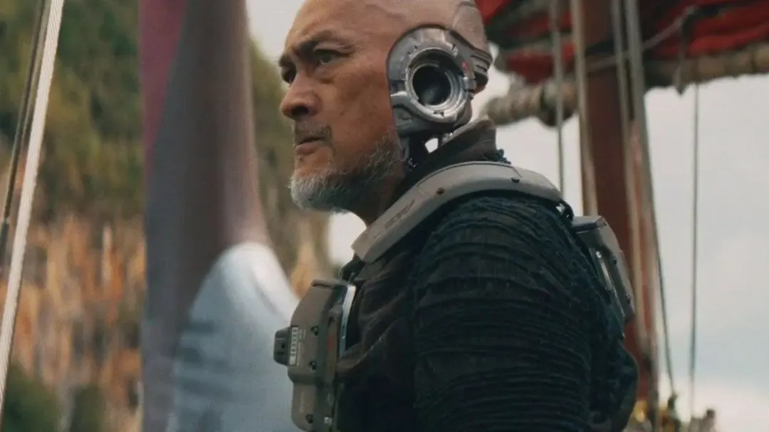 Homme chauve barbu portant une prothèse oculaire et une armure futuriste, dans un décor industriel.