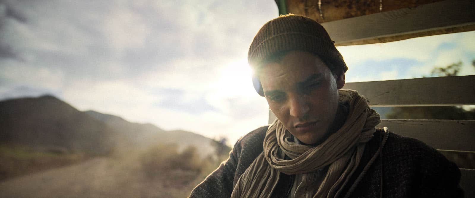 Un homme portant un bonnet et une écharpe, assis à l’arrière d’un véhicule face à des montagnes.