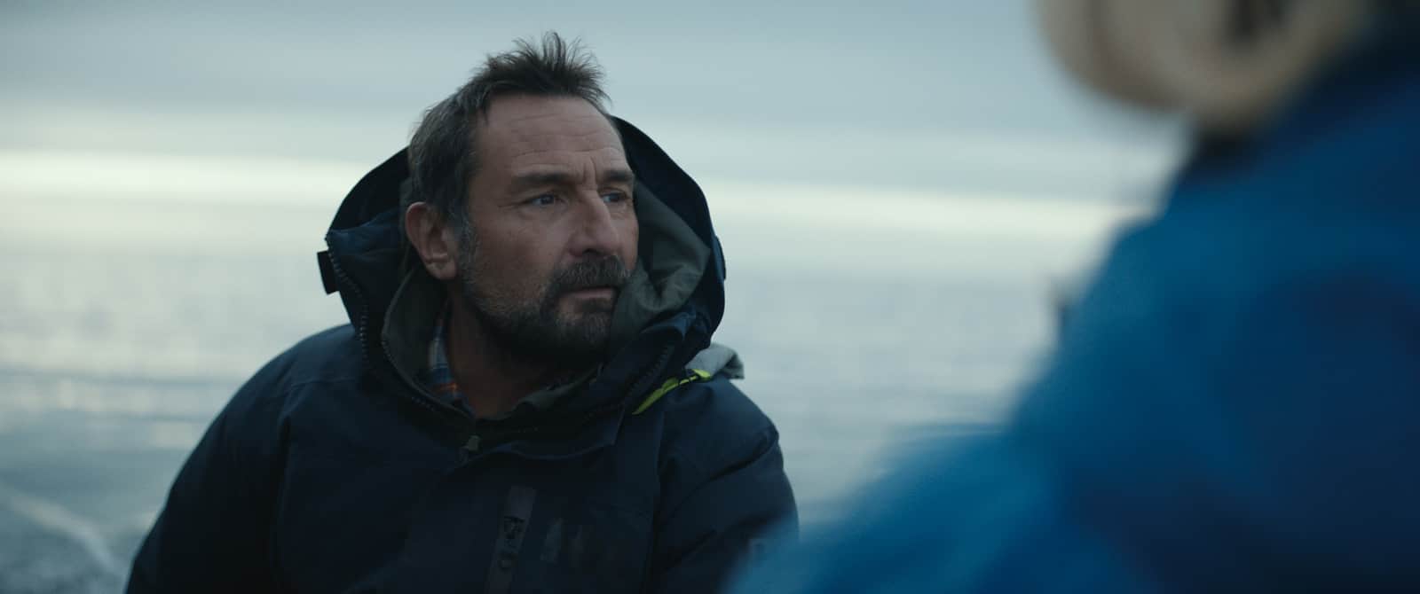 Un homme barbu en veste sombre regarde une personne en manteau bleu au bord de la mer.