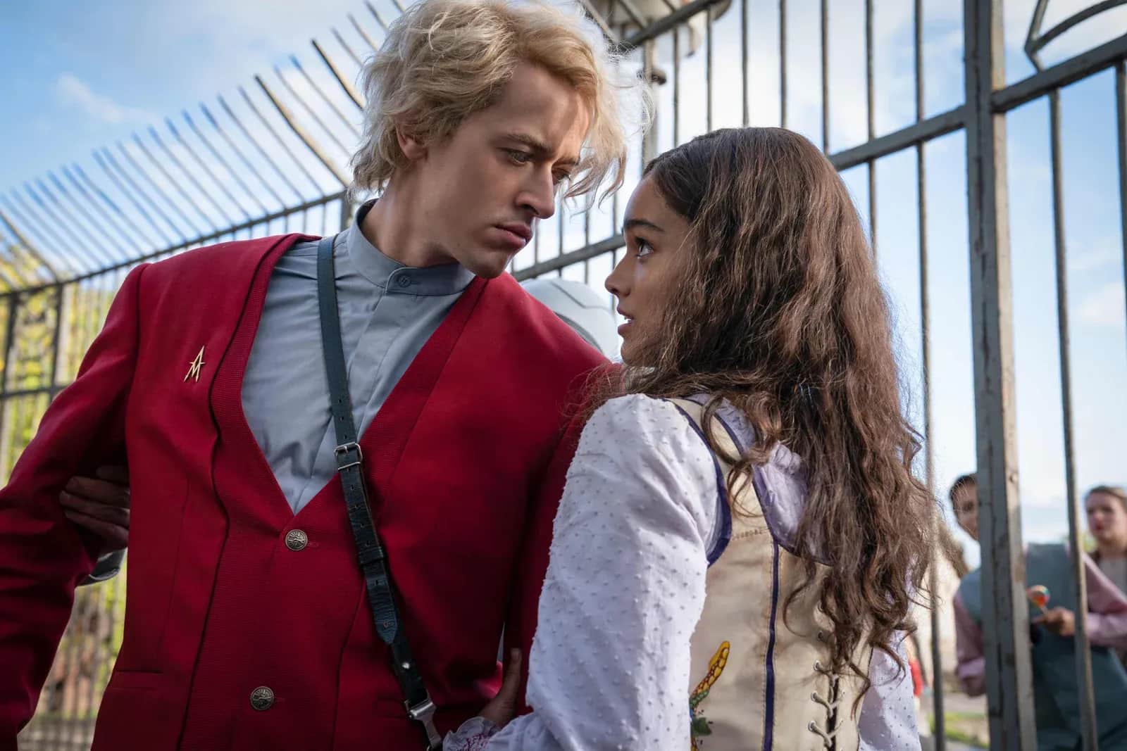 Deux jeunes gens se regardent dans une serre vitrée, l’homme portant une veste rouge et la femme une robe blanche.