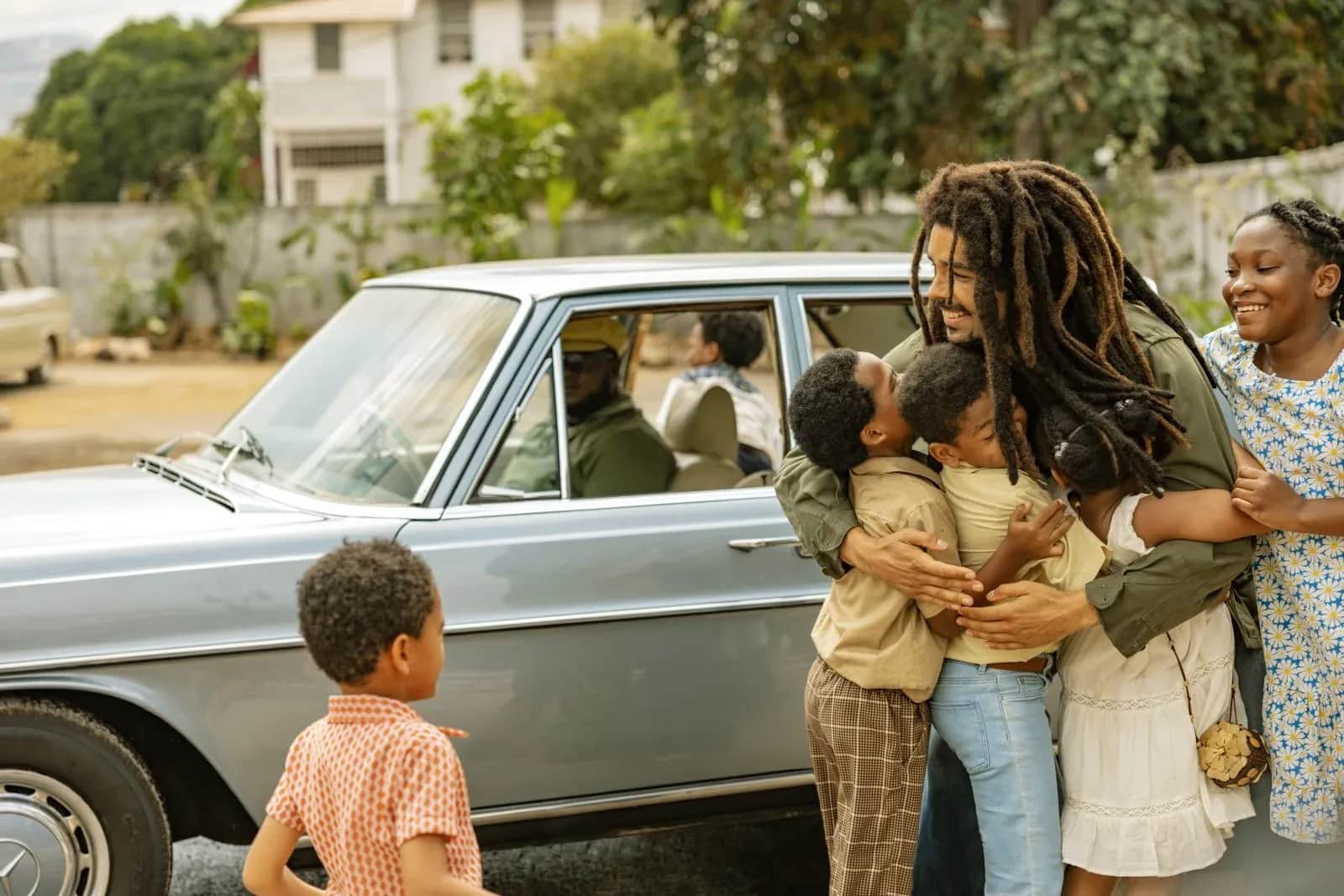 Un père enlace trois enfants près d'une voiture ancienne, tandis qu'une femme souriante se tient à leurs côtés.