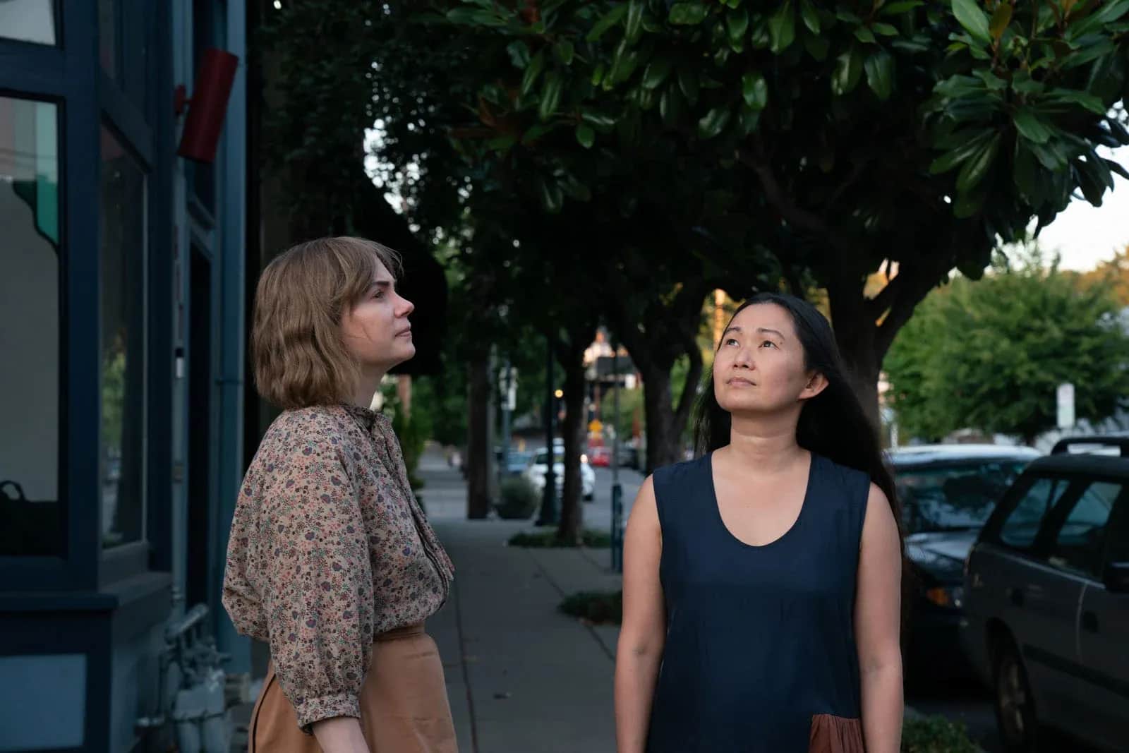Deux femmes regardent vers le ciel sur un trottoir bordé d’arbres et de voitures.