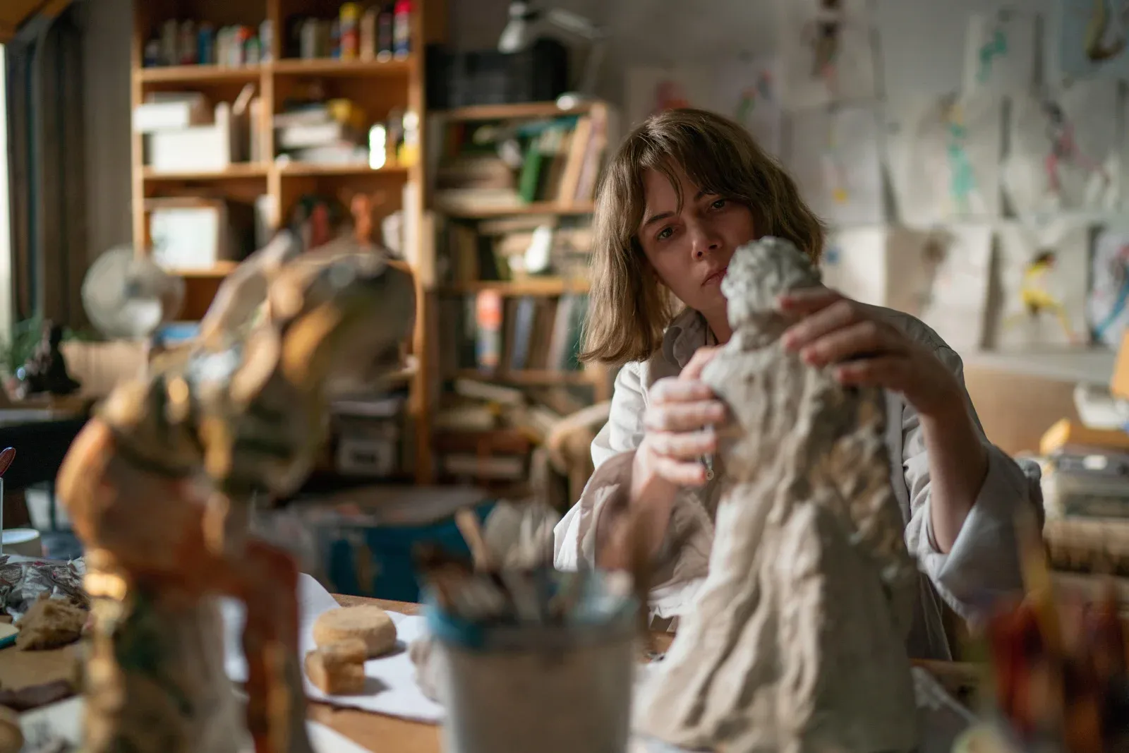 Femme sculptant une statue dans un atelier encombré de livres, de dessins et d’objets.