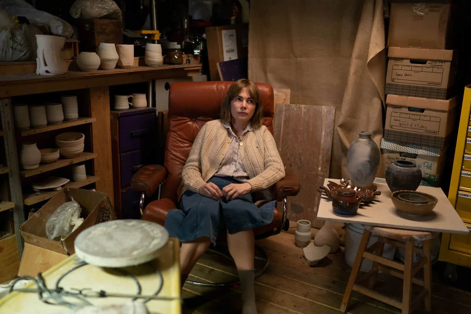 Une femme assise dans un fauteuil au milieu d’un atelier rempli de poteries et de cartons.