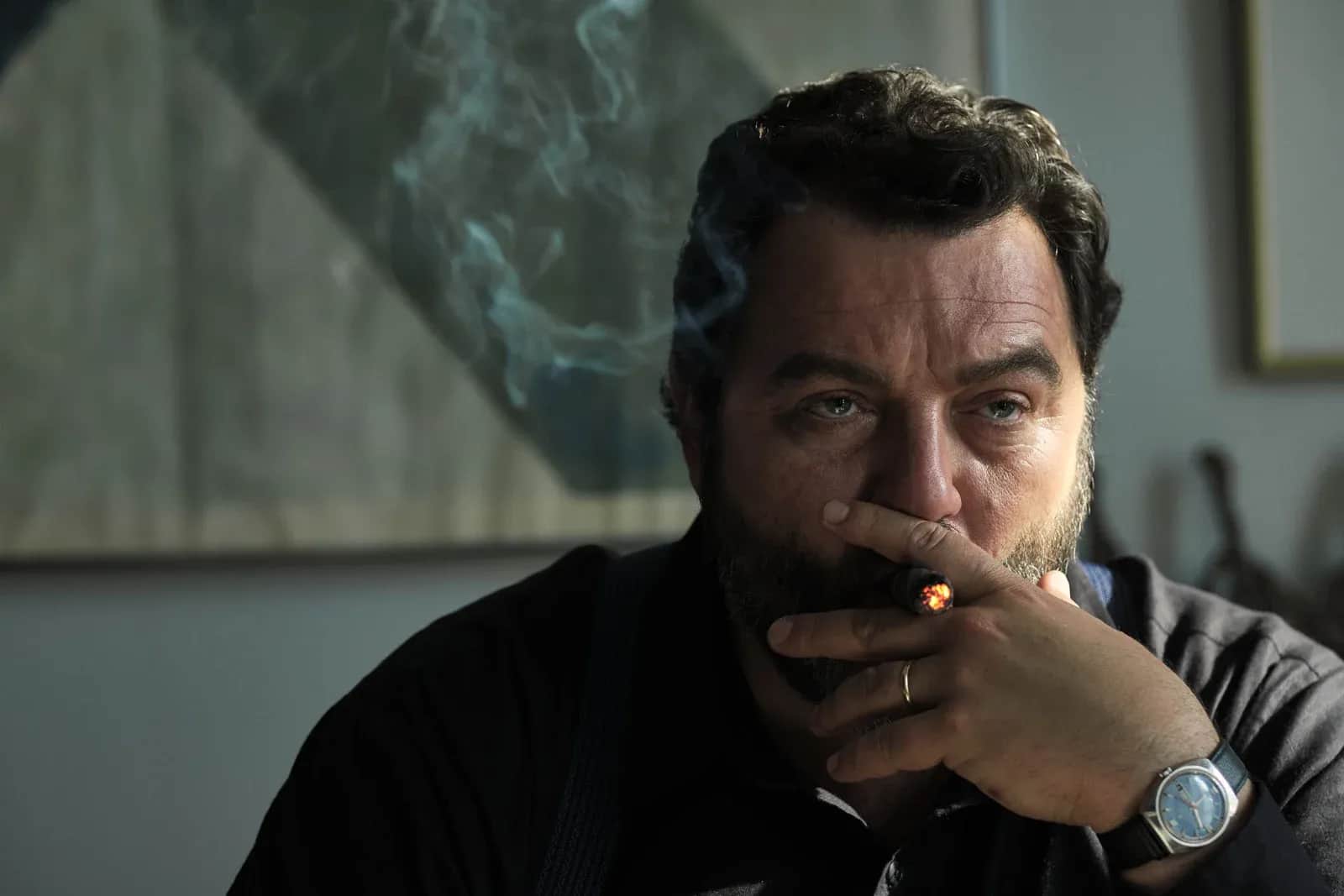 Homme barbu fumant un cigare, assis dans une pièce enfumée.