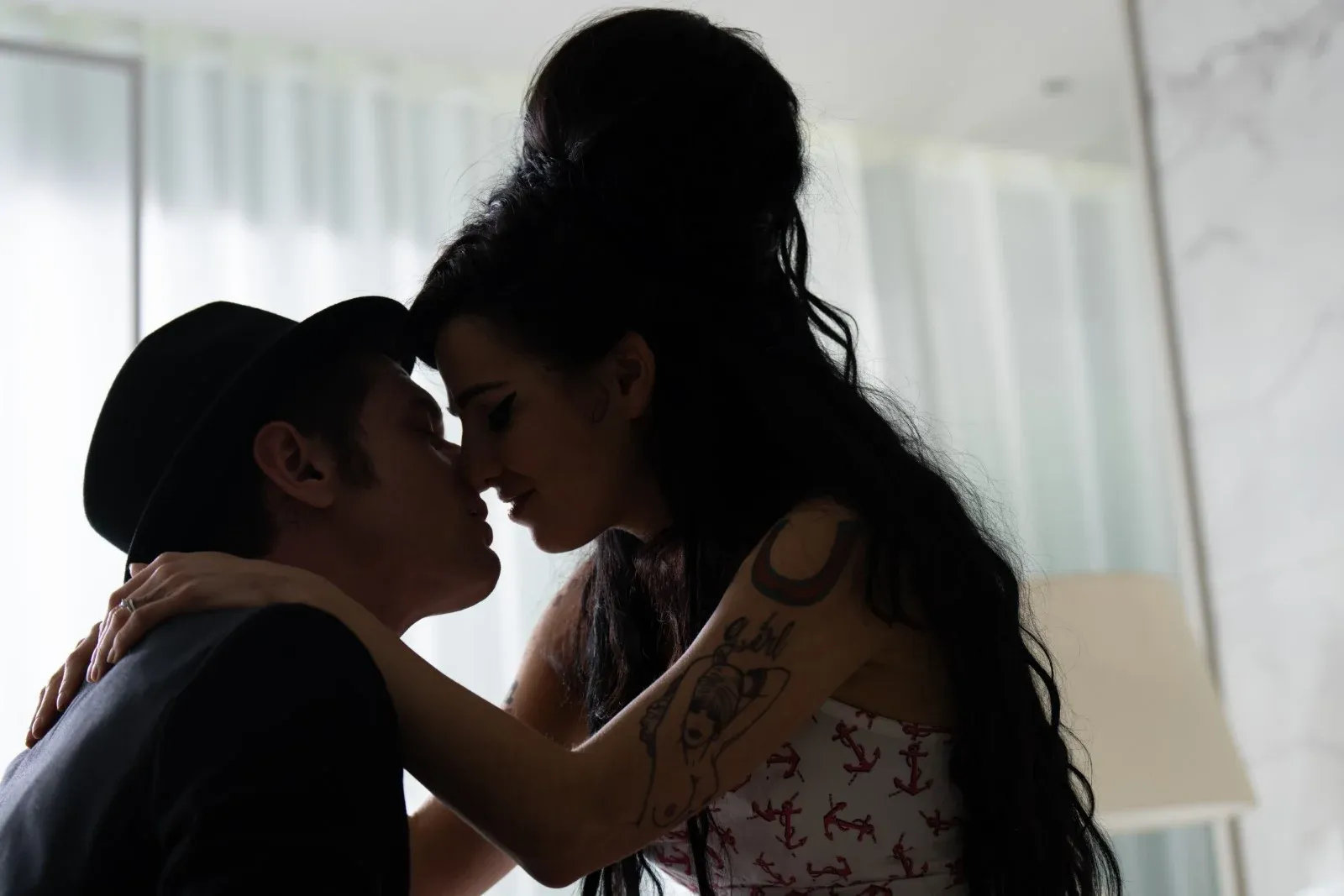 Un homme et une femme tatouée se rapprochent pour s’embrasser devant une fenêtre lumineuse