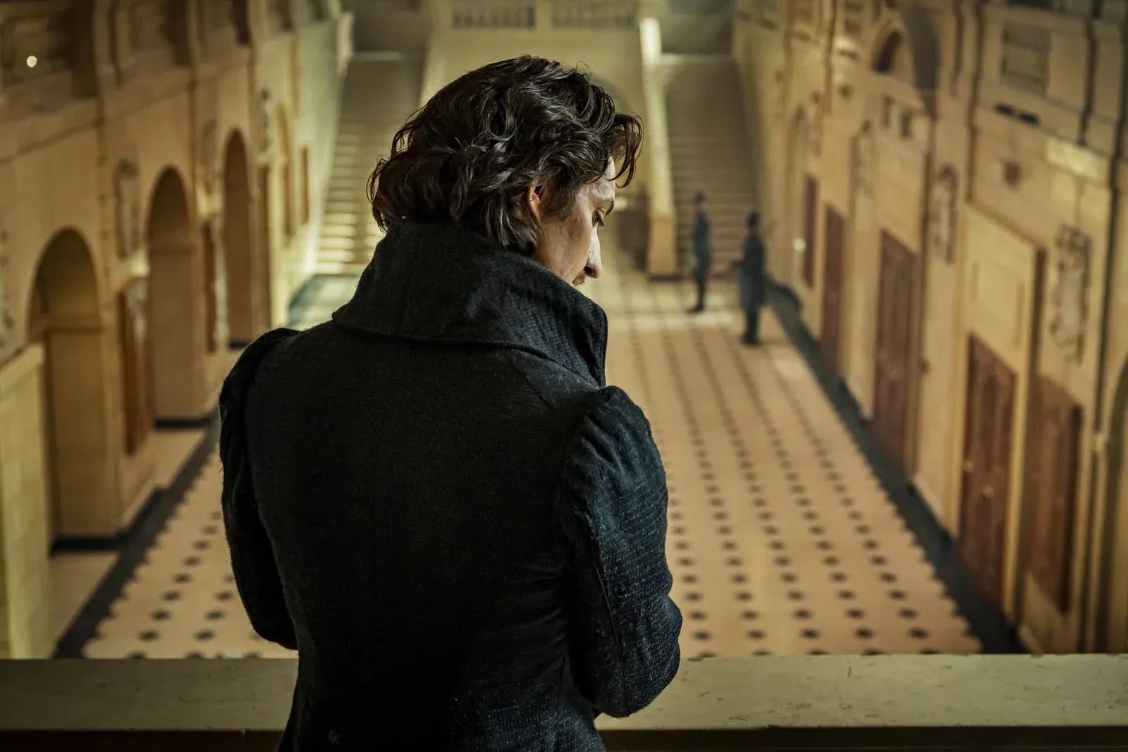 Personne de dos, vêtue de noir, se tient dans l’escalier d’un vaste bâtiment aux arcades.