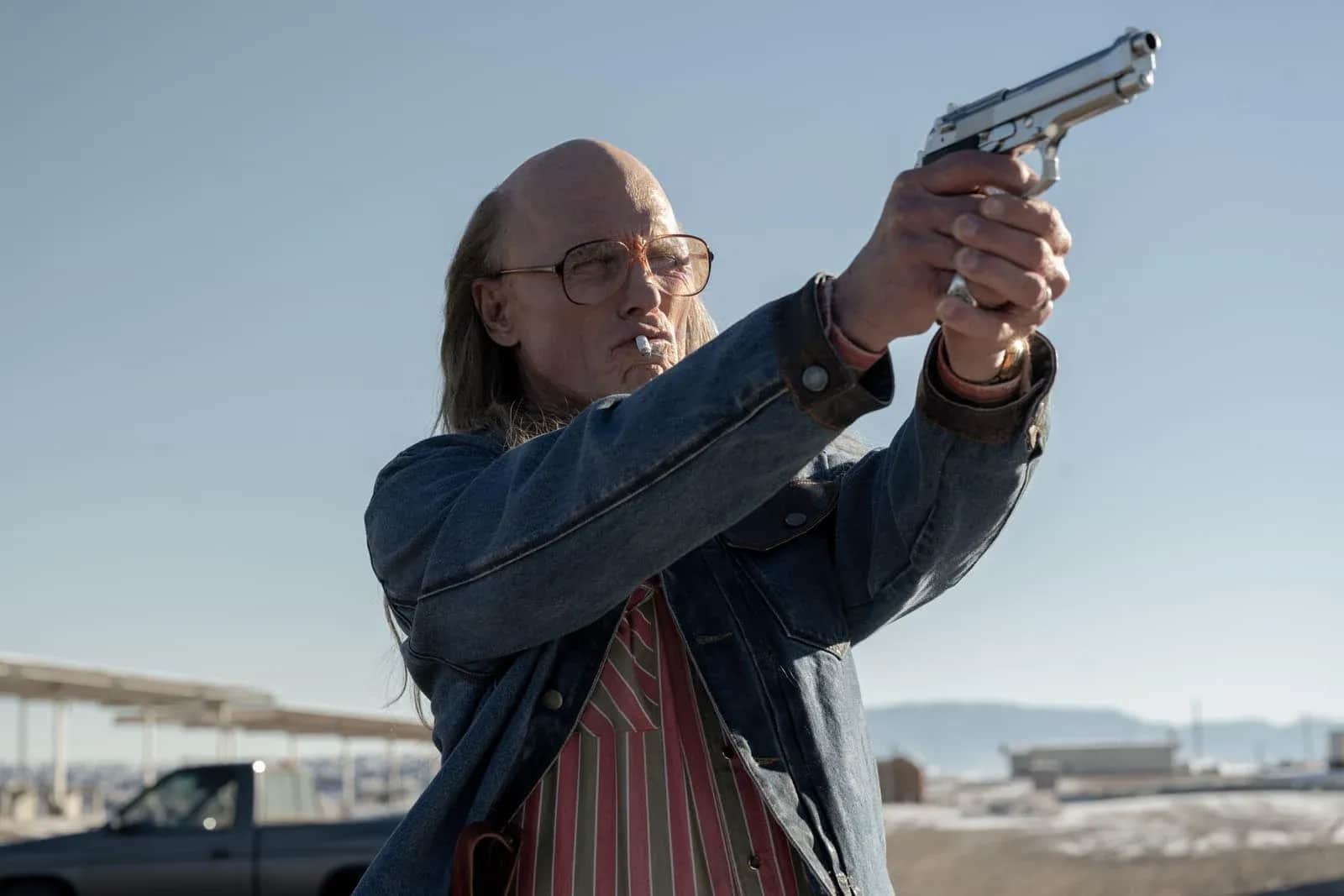 Un homme aux cheveux longs braque une arme de poing argentée vers le ciel, dans un paysage désertique.