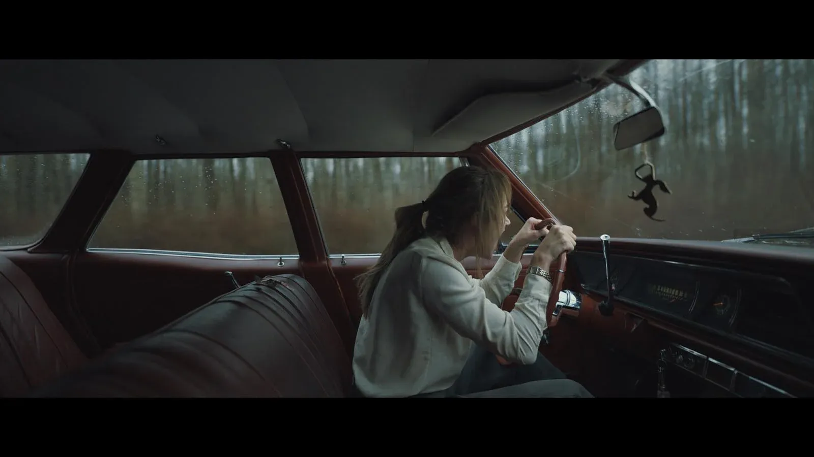 Une femme conduit une voiture ancienne à travers une forêt, vue depuis la banquette arrière.
