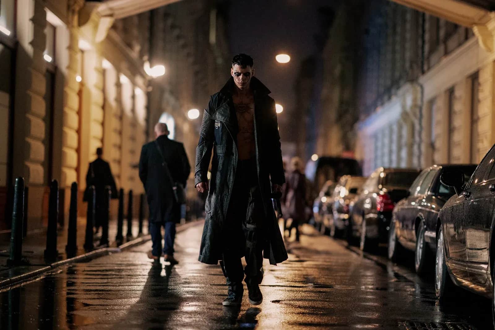 Homme en long manteau noir marchant dans une rue mouillée éclairée la nuit, entre des voitures garées.