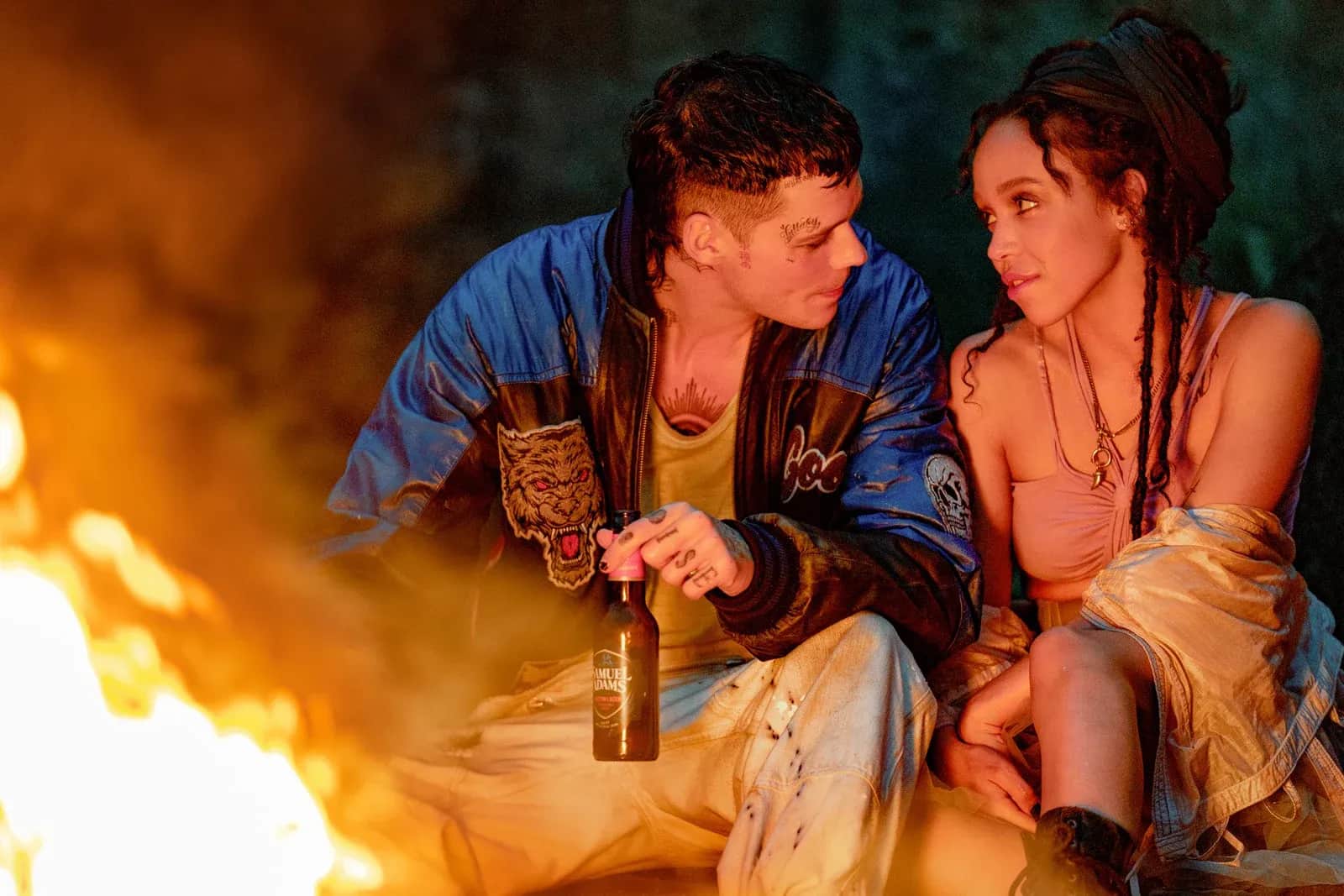 Un homme et une femme assis près d’un feu, se regardant, l’homme tenant une bouteille.