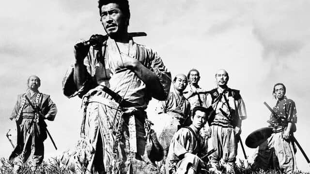 Un groupe de sept samouraïs en tenue traditionnelle, debout dans un champ, avec un homme au premier plan tenant une épée.