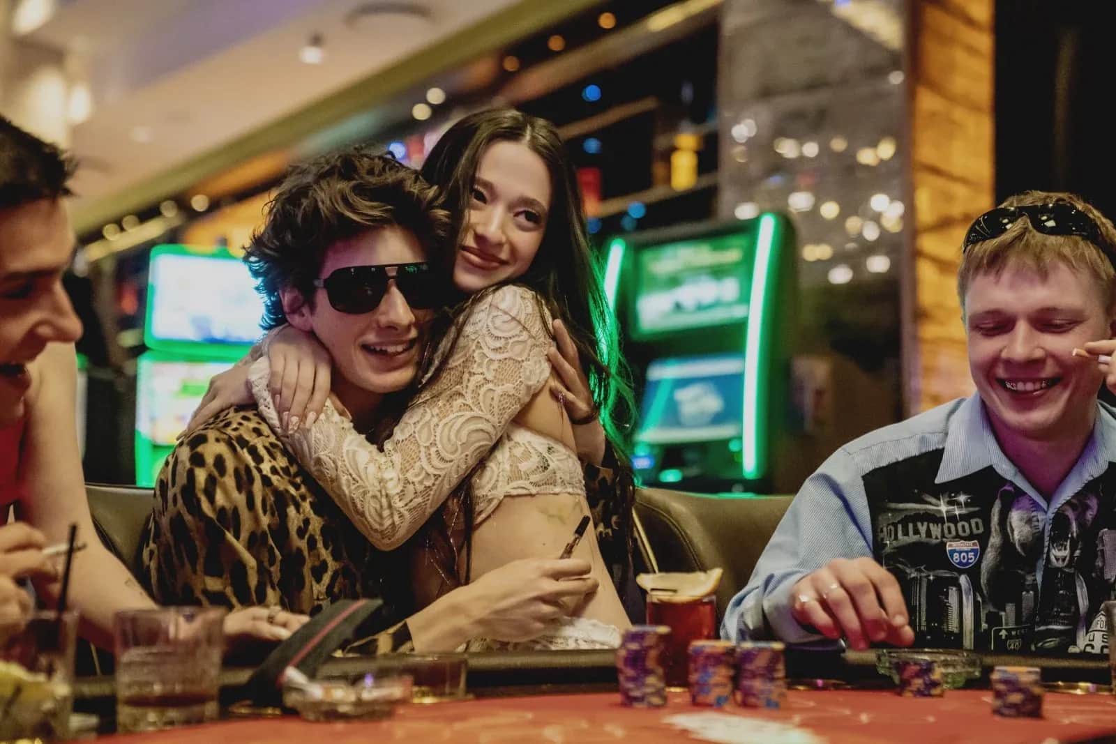 Un groupe de personnes attablées dans un casino, une femme enlace un homme portant des lunettes de soleil.