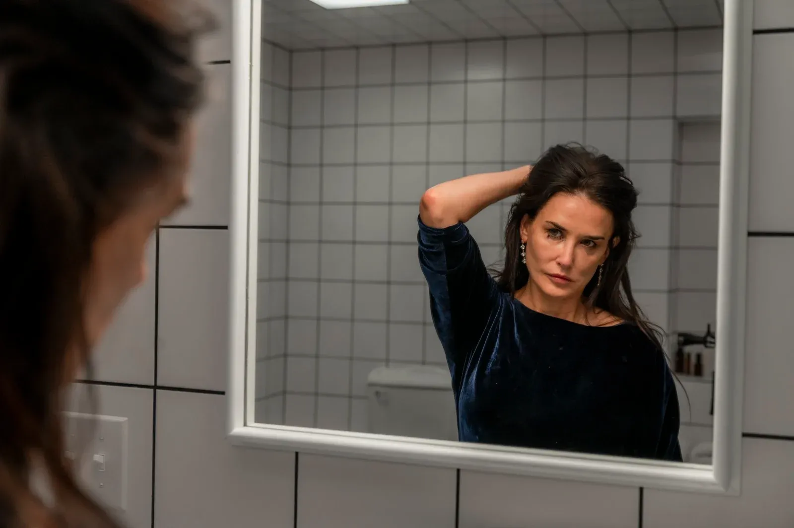 Une femme se regarde dans un miroir en remettant ses cheveux en place, dans une salle de bains carrelée
