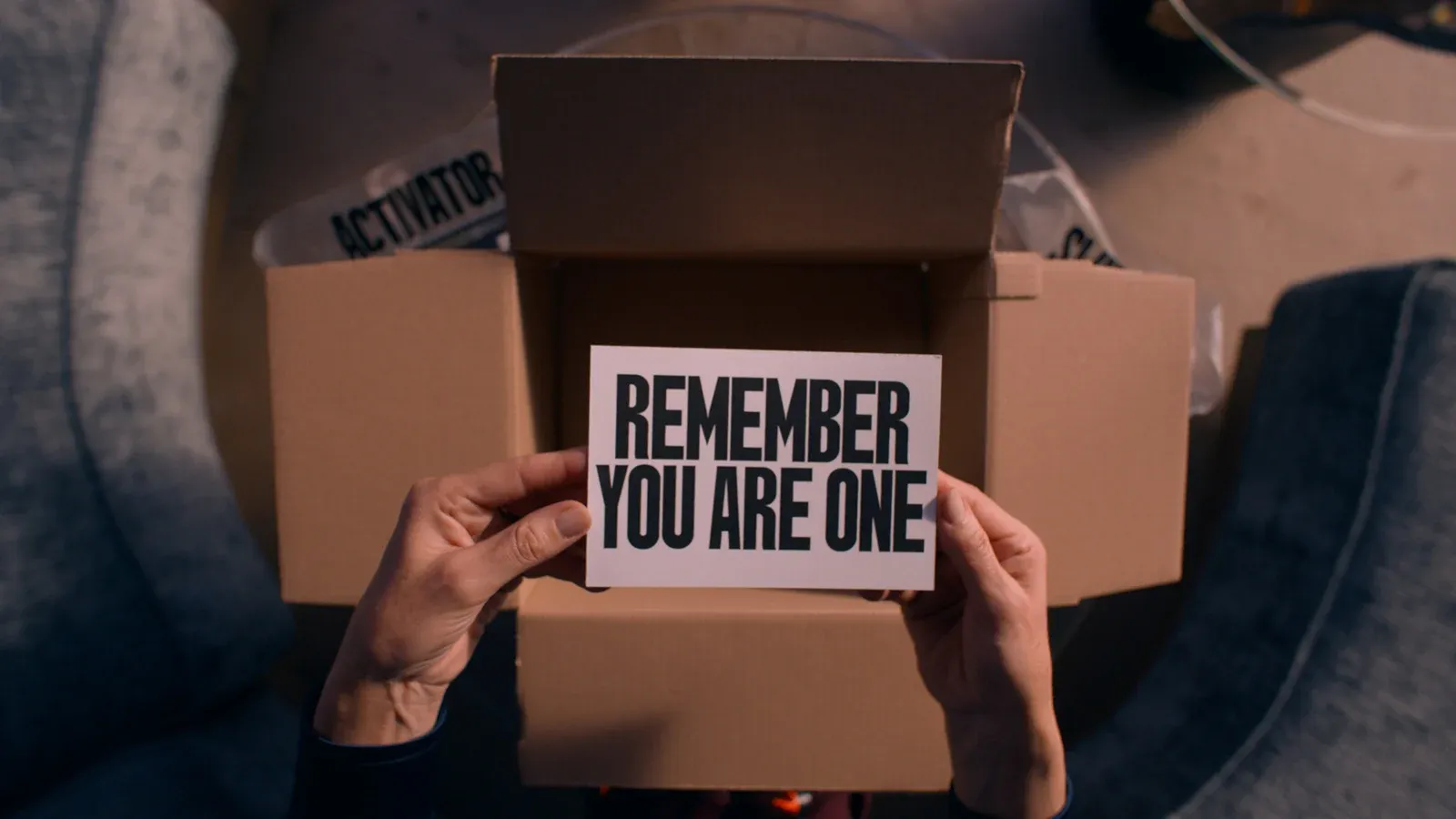 Deux mains tiennent un carton marqué « REMEMBER YOU ARE ONE » devant une boîte ouverte.
