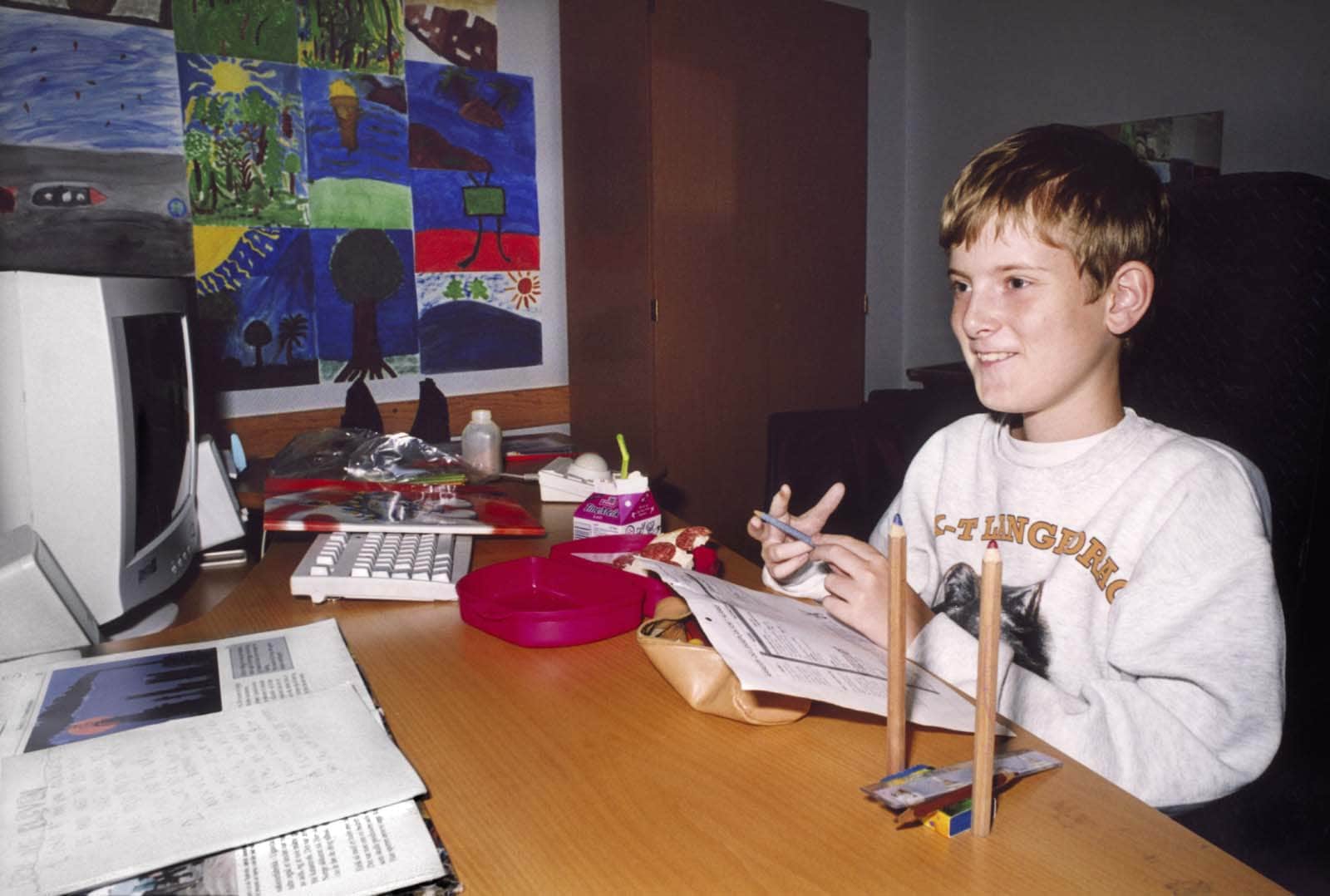 Garçon assis à un bureau, tenant un stylo et mangeant, devant un ordinateur et des dessins d’enfants.