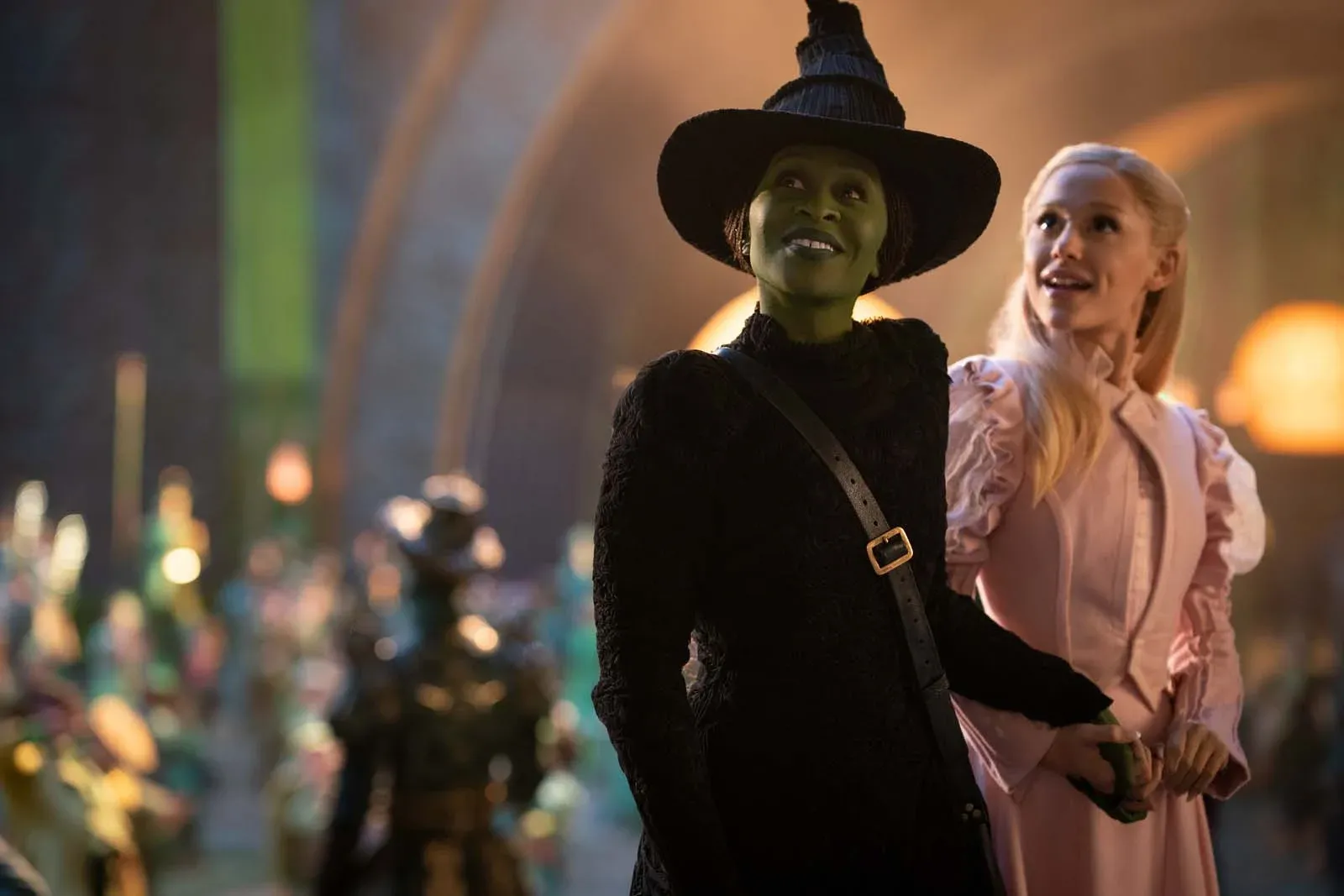 Une femme au visage vert et chapeau noir marche avec une femme blonde en robe rose dans une rue illuminée