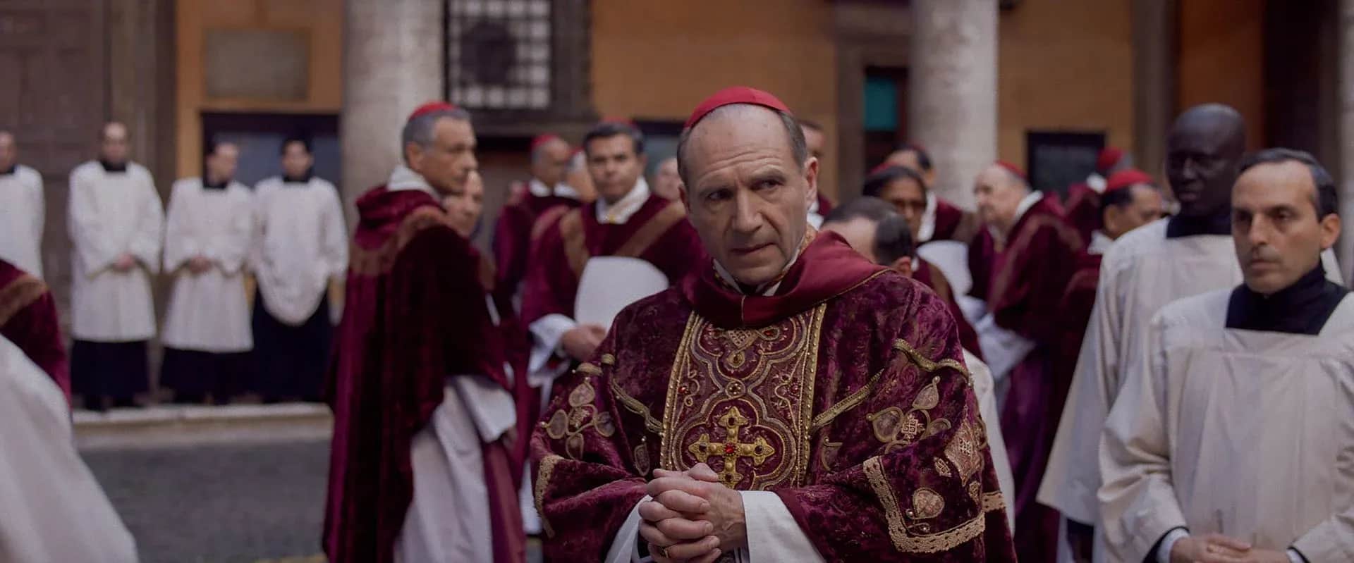 Un ecclésiastique en tenue liturgique pourpre, entouré de religieux dans une cour.