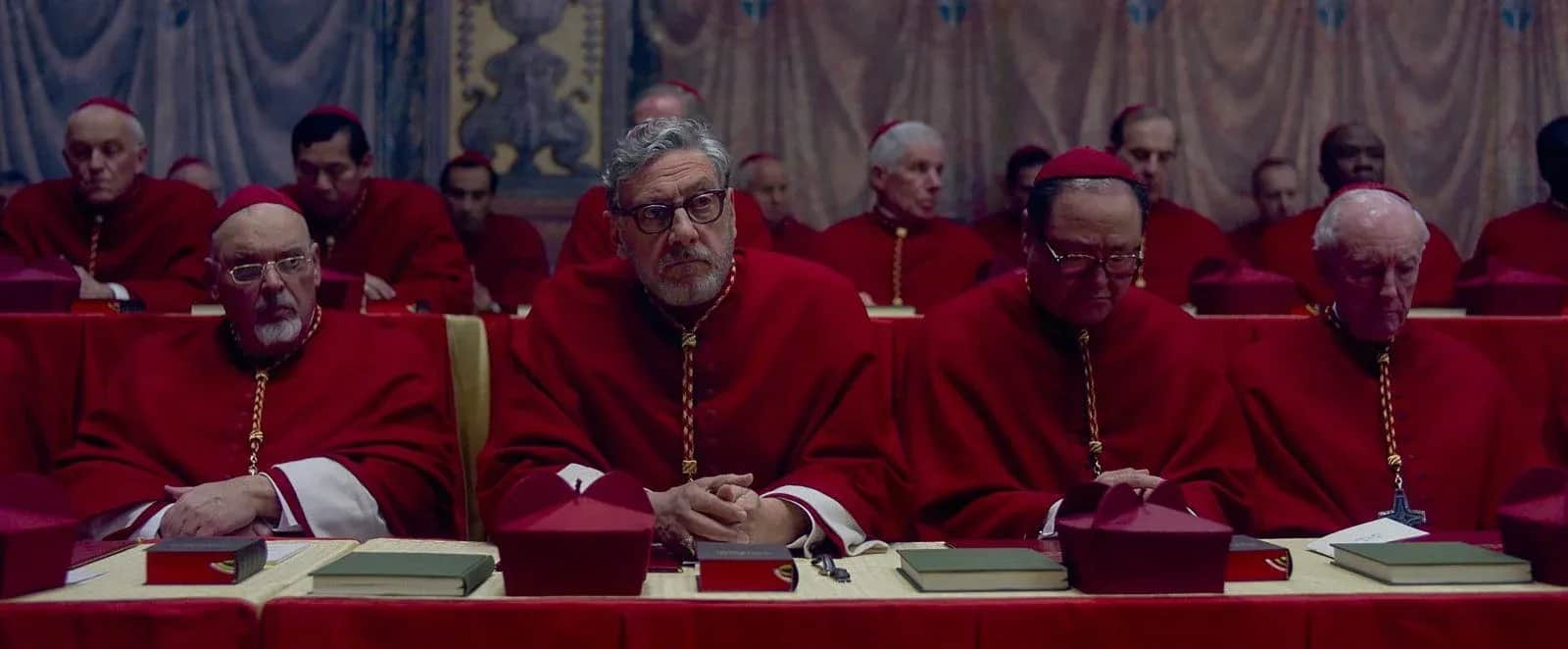 Un groupe de cardinaux en rouge assis autour d’une table dans une salle ornée de tentures