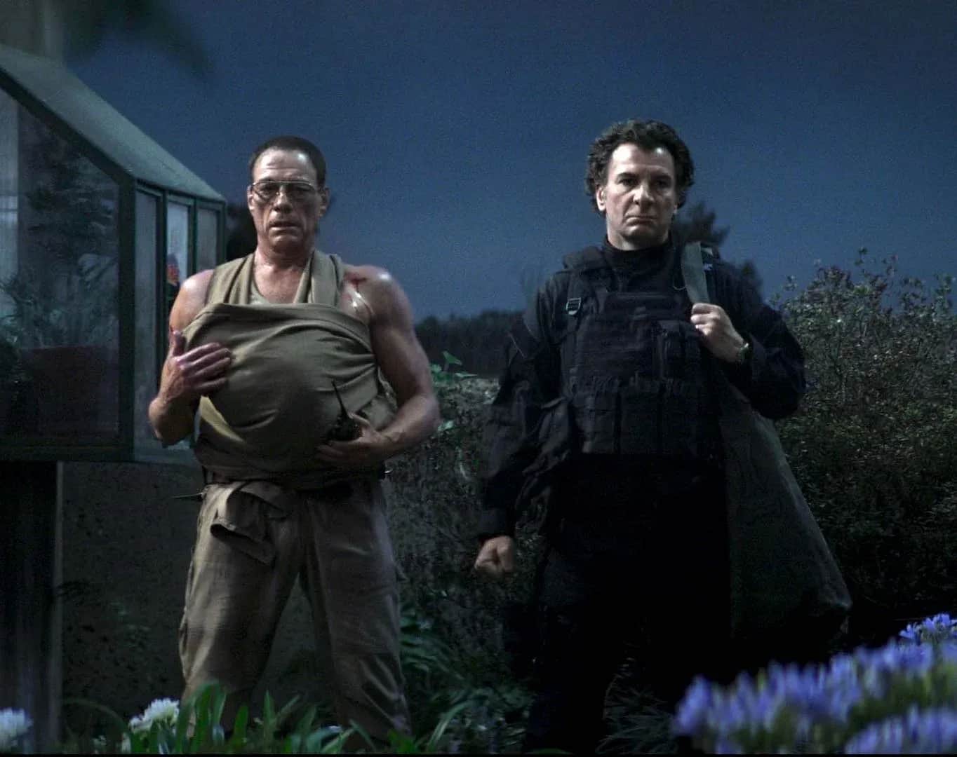 Deux hommes marchent de nuit dans un jardin, l’un portant un vêtement déchiré, l’autre vêtu en soldat.
