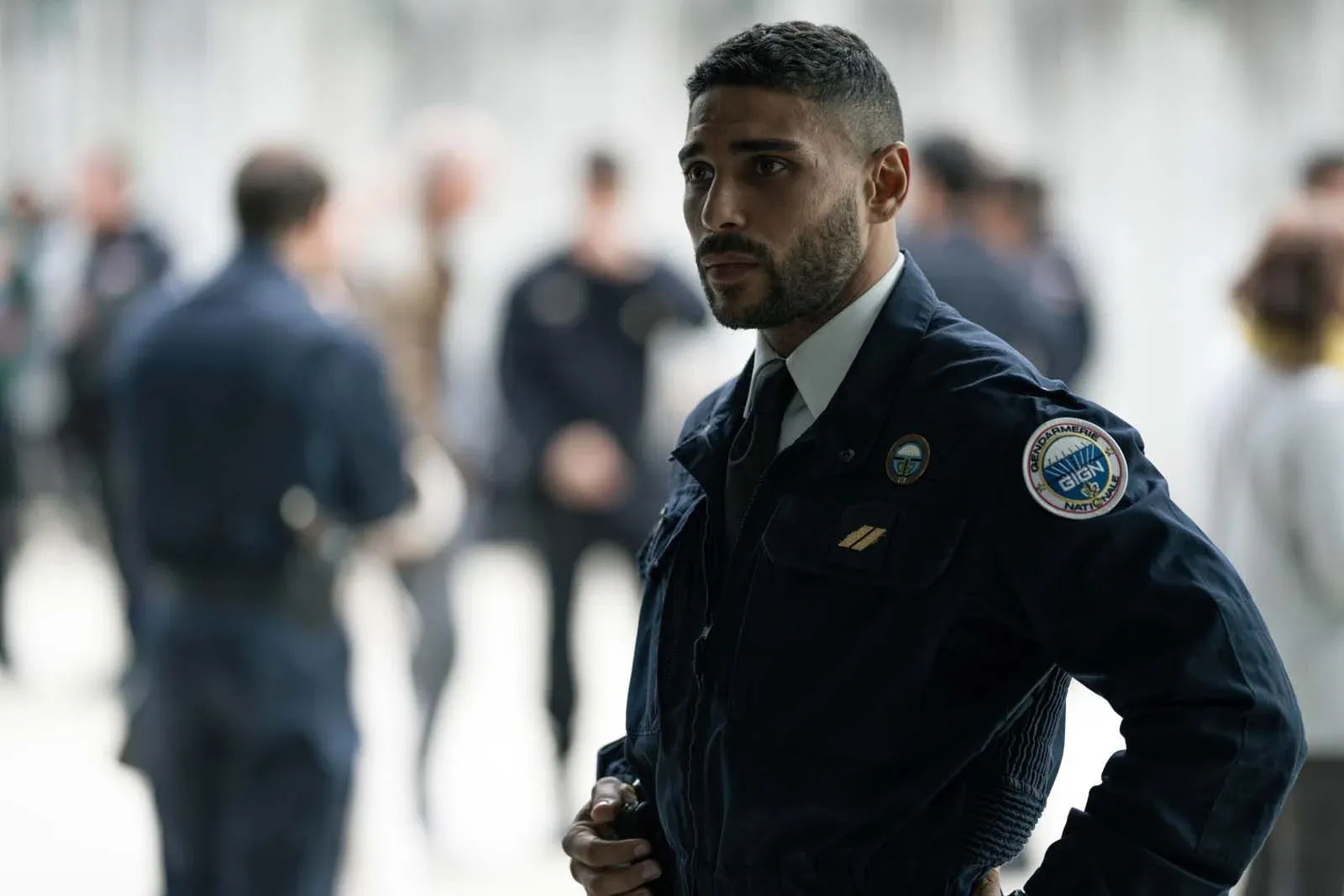 Un homme en uniforme de sécurité, debout dans un intérieur lumineux, avec plusieurs personnes floues en arrière-plan