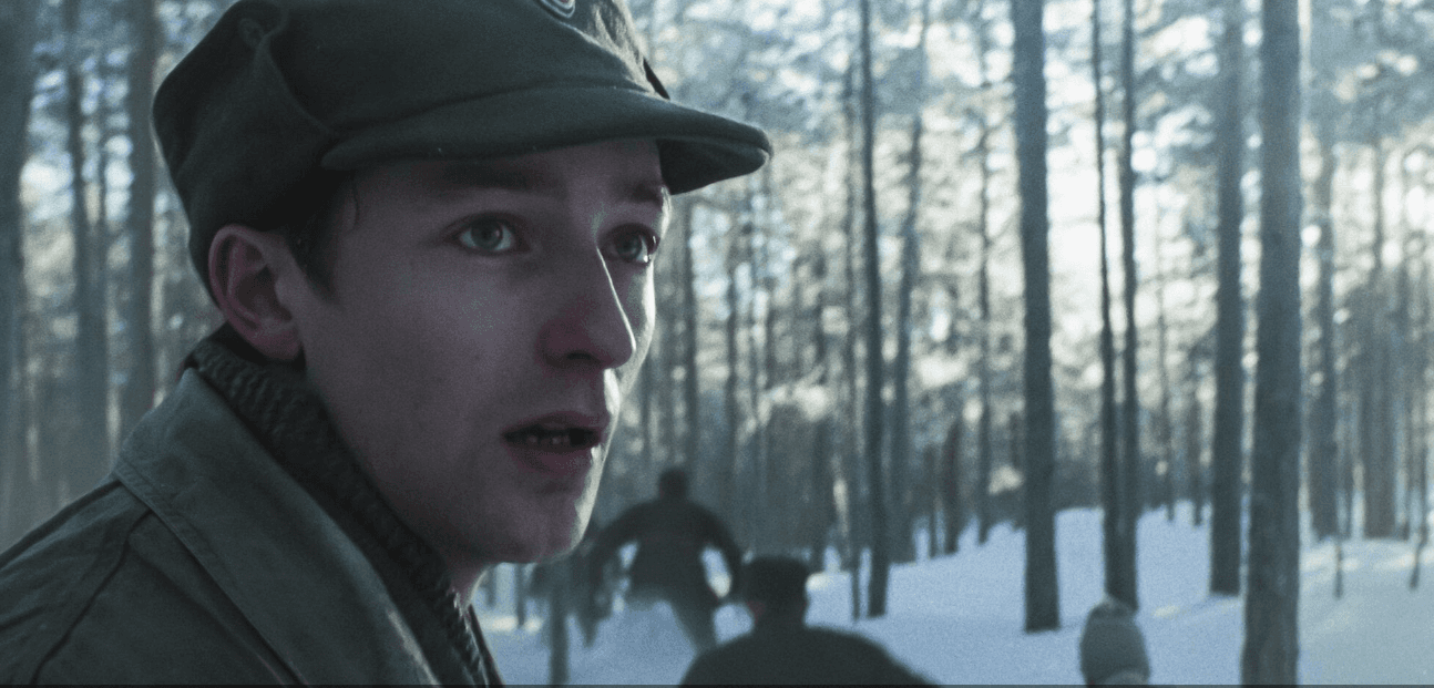 Jeune homme en casquette militaire dans une forêt enneigée, avec plusieurs personnes en arrière-plan.