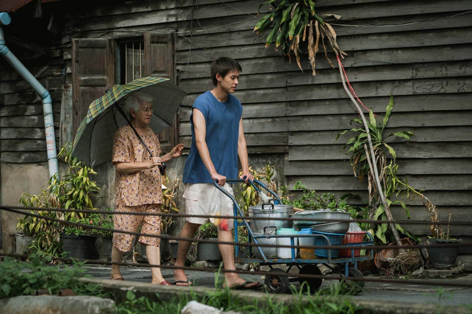 Un jeune homme pousse un chariot chargé d’ustensiles, accompagné d’une femme âgée sous un parapluie devant une maison en bois