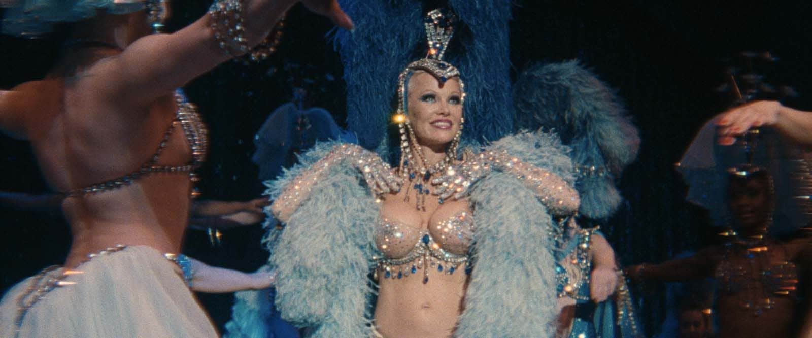 Une femme en costume scintillant et plumes bleues danse au milieu de danseurs dans un spectacle.