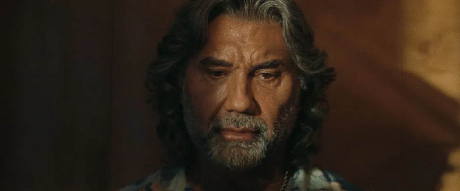 Homme barbu aux cheveux longs, les yeux baissés dans une pièce sombre.