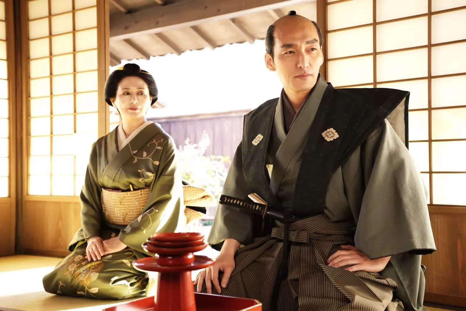 Un homme en tenue traditionnelle japonaise, assis devant une femme en kimono dans une maison japonaise.